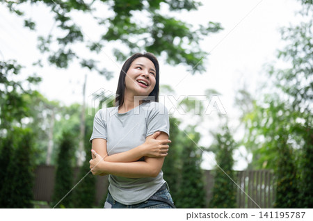 Beautiful Asian woman hugging herself and laughing happily in a backyard park. Beautiful Asian woman hugging herself and laughing happily in a backyard park. 141195787