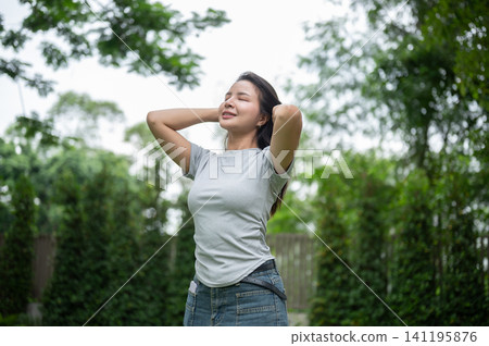 Relaxed Asian woman holding hands behind head and closing eyes in a garden. Relaxed Asian woman holding hands behind head and closing eyes in a garden. 141195876