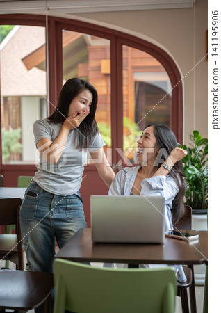 Asian women friends looking surprised while celebrating teamwork and success beside a laptop in cafe Asian women friends looking surprised while celebrating teamwork and success beside a laptop in cafe 141195906