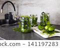 Cucumbers and herbs are arranged in jars on a kitchen counter. A sink is visible in the background.  141197283