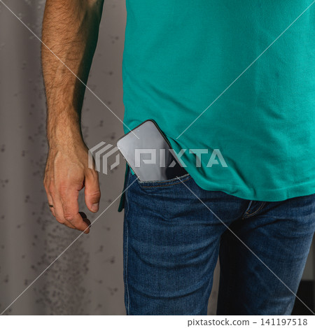 Man stands with a smartphone in his jeans pocket against a simple background 141197518
