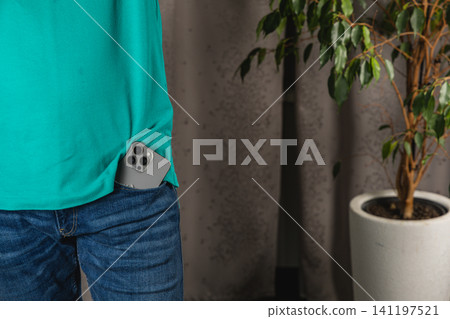 Man stands with smartphone in jeans pocket next to a plant Man stands with smartphone in jeans pocket next to a plant 141197521