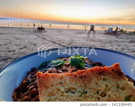 Bolognese pasta with garlic toast, enjoyed against the backdrop of a beautiful sunset at Seminyak Beach in Bali. Bolognese pasta with garlic toast, enjoyed against the backdrop of a beautiful sunset at Seminyak Beach in Bali. 141198445
