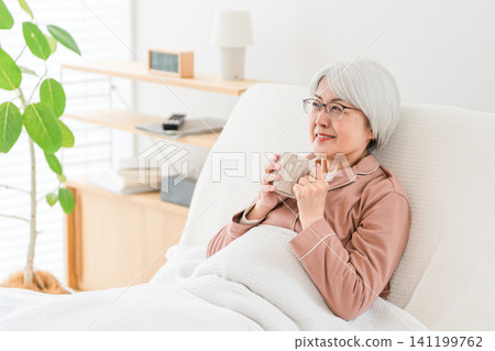 一位頭髮花白的老婦人,穿著睡衣,從臥室或醫院病房的護理床上起身後,正在喝水。 一位頭髮花白的老婦人,穿著睡衣,從臥室或醫院病房的護理床上起身後,正在喝水。 141199762