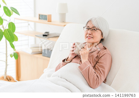 A senior woman with gray hair, dressed in pajamas, hydrates herself after getting up from a nursing bed in a bedroom or hospital room. A senior woman with gray hair, dressed in pajamas, hydrates herself after getting up from a nursing bed in a bedroom or hospital room. 141199763
