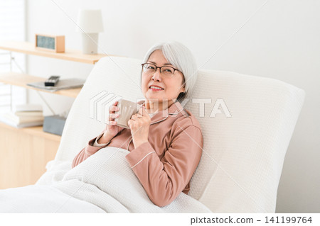 A senior woman with gray hair, dressed in pajamas, hydrates herself after getting up from a nursing bed in a bedroom or hospital room. A senior woman with gray hair, dressed in pajamas, hydrates herself after getting up from a nursing bed in a bedroom or hospital room. 141199764