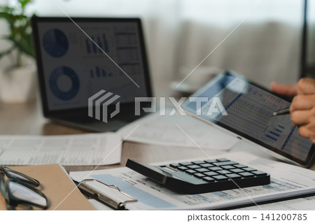 Businessman hand reviews financial data on a tablet Calculator rests on printed reports dashboard, showing document analysis and accounting report checking for business workflow. Businessman hand reviews financial data on a tablet Calculator rests on printed reports dashboard, showing document analysis and accounting report checking for business workflow. 141200785