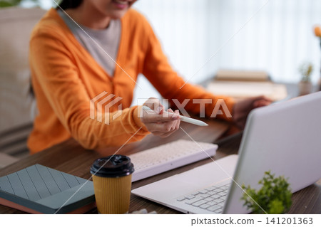 Adult Asian woman writing notes with stylus pen while studying online with laptop at home desk 141201363