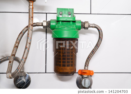 Dirty water filter mesh housing connected with rusty braided hoses on a white tiled wall backdrop. Dirty water filter mesh housing connected with rusty braided hoses on a white tiled wall backdrop. 141204703