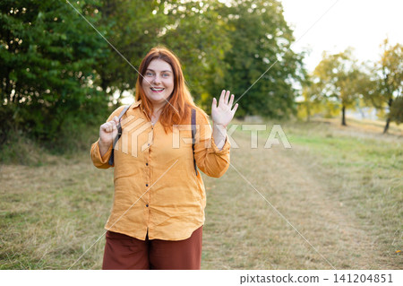 Smiling Red Haired Woman Backpacker Waving Hand in Green Forest Smiling Red Haired Woman Backpacker Waving Hand in Green Forest 141204851