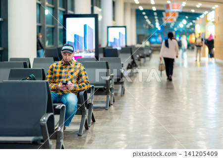 Wide shot of adult Caucasian man in cap and jacket using smartphone in airport waiting area sitting in chair. Copy space. Concept of travel and trip 141205409