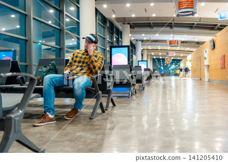 Wide shot of adult Caucasian man in cap and jacket using smartphone in airport waiting area sitting in chair. Low angle view. Copy space. Concept of trip and travel Wide shot of adult Caucasian man in cap and jacket using smartphone in airport waiting area sitting in chair. Low angle view. Copy space. Concept of trip and travel 141205410