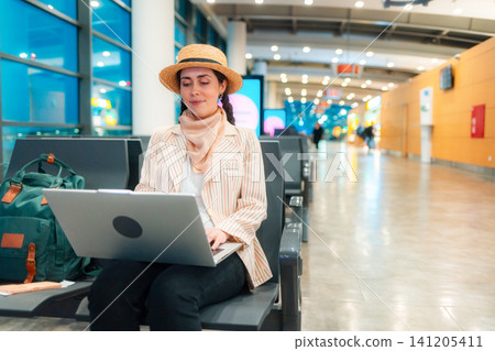 Young Caucasian elegant happy woman is sitting on a chair in waiting room of airport and working on a laptop. Low angle view. Copy space. Concept of remote work and travel Young Caucasian elegant happy woman is sitting on a chair in waiting room of airport and working on a laptop. Low angle view. Copy space. Concept of remote work and travel 141205411