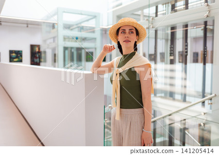 Mid shot of young Caucasian elegant woman in suit and straw hat posing in the modern art gallery. Concept of cultural education and Museum day Mid shot of young Caucasian elegant woman in suit and straw hat posing in the modern art gallery. Concept of cultural education and Museum day 141205414