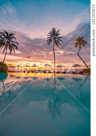Vertical view of resort infinity pool at dusk with palm silhouettes and umbrella reflections. Purple and orange twilight sky mirrored in calm pool water by tropical beach. Moody evening luxury scene Vertical view of resort infinity pool at dusk with palm silhouettes and umbrella reflections. Purple and orange twilight sky mirrored in calm pool water by tropical beach. Moody evening luxury scene 141205813