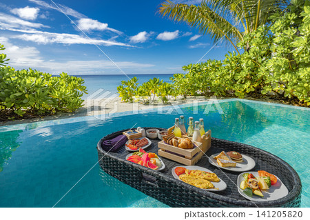 Floating breakfast basket with fresh food and drinks in private pool by the beach. Tropical morning meal on water, lush plants and turquoise sea behind. Trendy luxury vacation experience for resort  141205820