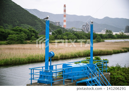 Under a cloudy, rainy sky, birds perch on a blue sluice gate, with factories, chimneys, and mountain ranges visible in the background. Under a cloudy, rainy sky, birds perch on a blue sluice gate, with factories, chimneys, and mountain ranges visible in the background. 141206291