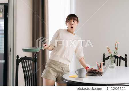 Surprised young asian woman expressing panic holding green frying pan in bright dining room during morning daily lifestyle routine near table with laptop computer and coffee mug 141206311