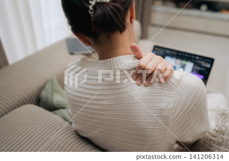 Exhausted asian woman sitting on comfortable sofa experiencing severe shoulder pain during stressful laptop work representing difficult remote office lifestyle with intense uncomfortable feeling Exhausted asian woman sitting on comfortable sofa experiencing severe shoulder pain during stressful laptop work representing difficult remote office lifestyle with intense uncomfortable feeling 141206314