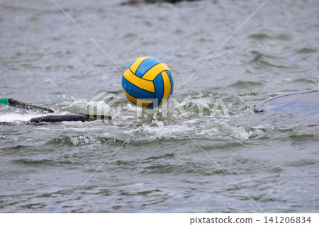 Colorful blue and yellow ball splashes in water during a canoe polo match, with paddles partially visible and ripples forming around the ball in a competitive setting Colorful blue and yellow ball splashes in water during a canoe polo match, with paddles partially visible and ripples forming around the ball in a competitive setting 141206834