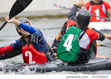 Two athletes engaged in canoe polo match, one wearing red jersey with number 2 and the other in green jersey with number 4, paddling in water with splashes visible Two athletes engaged in canoe polo match, one wearing red jersey with number 2 and the other in green jersey with number 4, paddling in water with splashes visible 141206842