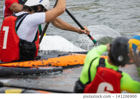 Canoe polo players in colorful gear maneuvering kayaks on water, with splashes visible as they engage in a competitive match during a sporting event Canoe polo players in colorful gear maneuvering kayaks on water, with splashes visible as they engage in a competitive match during a sporting event 141206900