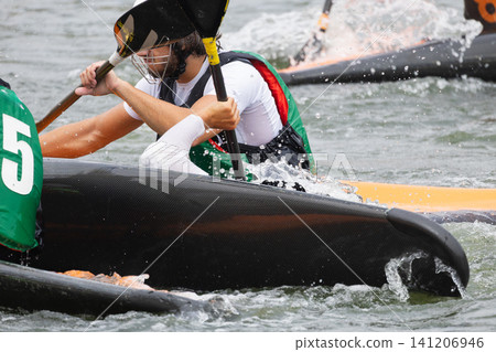 Male athlete in a green canoe polo jersey paddles vigorously in a water sport competition, with splashes and other canoes visible in the background Male athlete in a green canoe polo jersey paddles vigorously in a water sport competition, with splashes and other canoes visible in the background 141206946