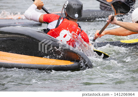 Male canoe polo player in red jersey paddles vigorously in water, splashes visible as he competes with other players in the background during a match Male canoe polo player in red jersey paddles vigorously in water, splashes visible as he competes with other players in the background during a match 141206964