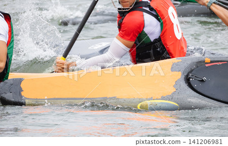 Canoe polo players in red and green jerseys maneuvering their kayaks on a water surface, with splashes and paddles visible during an intense match Canoe polo players in red and green jerseys maneuvering their kayaks on a water surface, with splashes and paddles visible during an intense match 141206981