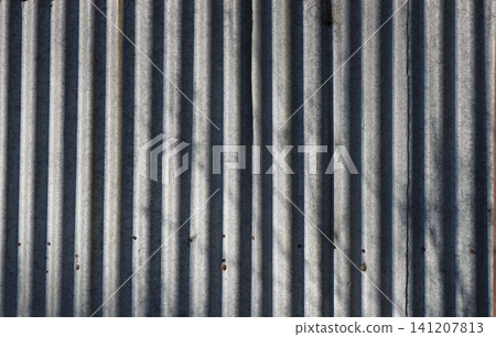The texture of old corrugated metal sheets with vertical relief. The galvanized gray surface is covered with small spots, traces of fasteners, and soft shadows from trees. Textured metal background The texture of old corrugated metal sheets with vertical relief. The galvanized gray surface is covered with small spots, traces of fasteners, and soft shadows from trees. Textured metal background 141207813