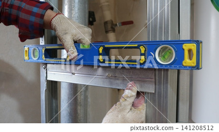 A worker checking the accuracy of the installation of galvanized metal profiles using a blue spirit level. The frame of the future wall is visible in the frame against the backdrop of utility lines. A worker checking the accuracy of the installation of galvanized metal profiles using a blue spirit level. The frame of the future wall is visible in the frame against the backdrop of utility lines. 141208512