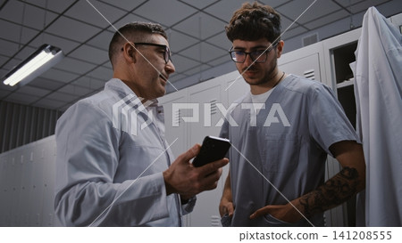Hospital Mentor and Intern Surgeon Reviewing Scientific Research on Smartphone in Changing Room Hospital Mentor and Intern Surgeon Reviewing Scientific Research on Smartphone in Changing Room 141208555