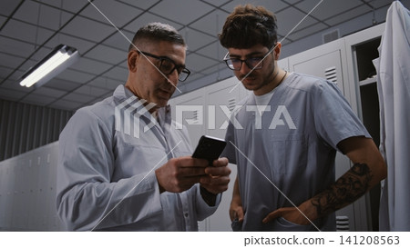 Hospital Mentor and Intern Surgeon Reviewing Scientific Research on Smartphone in Changing Room Hospital Mentor and Intern Surgeon Reviewing Scientific Research on Smartphone in Changing Room 141208563