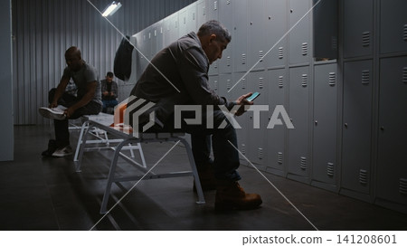 Factory Worker Sitting on Bench and Scrolling Social Media on Smartphone in Locker Room 141208601
