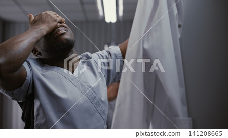 Male Doctor Undressing at Locker, Rubbing Face after Long Hospital Shift 141208665