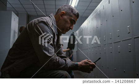 Factory Worker Taking Off Hard Hat, Checking Wristwatch and Texting on Smartphone in Locker Room Factory Worker Taking Off Hard Hat, Checking Wristwatch and Texting on Smartphone in Locker Room 141208667