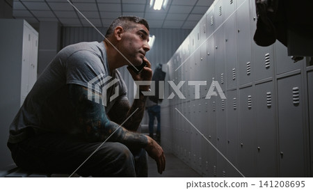 Tattooed Worker Making Phone Call on Smartphone in Factory Locker Room While Colleagues Change Tattooed Worker Making Phone Call on Smartphone in Factory Locker Room While Colleagues Change 141208695