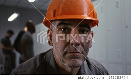 Serious Manual Laborer in Orange Hard Hat Sitting and Staring at Camera in Dressing Area Serious Manual Laborer in Orange Hard Hat Sitting and Staring at Camera in Dressing Area 141208734