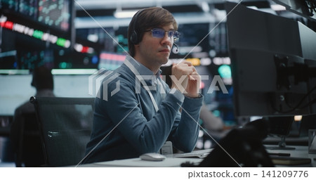 Young Male Financial Analyst in Glasses and Headset Sits at Modern Workstation. Young Male Financial Analyst in Glasses and Headset Sits at Modern Workstation. 141209776
