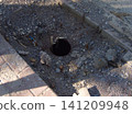 A small, dark circular hole in the ground at a construction site, surrounded by gravel, loose soil, and debris. Exposed electrical wires protrude from the opening near a paved curb 141209948