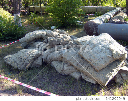 A pile of old, discarded mineral wool insulation lies on the grass near large industrial pipes. The area is cordoned off with safety tape, likely during maintenance work on the heating system A pile of old, discarded mineral wool insulation lies on the grass near large industrial pipes. The area is cordoned off with safety tape, likely during maintenance work on the heating system 141209949