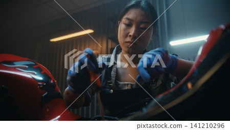 Focused Young Woman in Protective Gloves Repairs Motorcycle in Modern Garage Focused Young Woman in Protective Gloves Repairs Motorcycle in Modern Garage 141210296