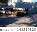 A stack of large, weathered metal pipes with visible rust lies on the pavement near a fenced industrial area. The heavy cylindrical components appear worn, awaiting transport or installation at a site 141210323