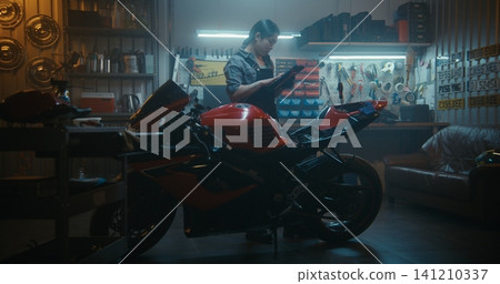 Woman Using Tablet to Check Motorcycle Electronics and Battery Wiring in Garage Woman Using Tablet to Check Motorcycle Electronics and Battery Wiring in Garage 141210337