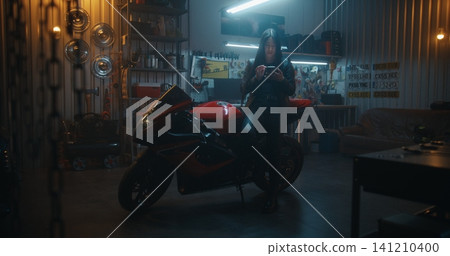 Female Biker With Long Black Hair Leans on Sport bike While Using Smartphone 141210400