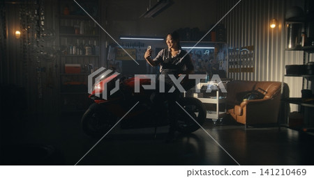 Female Rider Rests Against Sleek Red Sport bike Inside Modern Garage, Chatting Via Video Call Female Rider Rests Against Sleek Red Sport bike Inside Modern Garage, Chatting Via Video Call 141210469