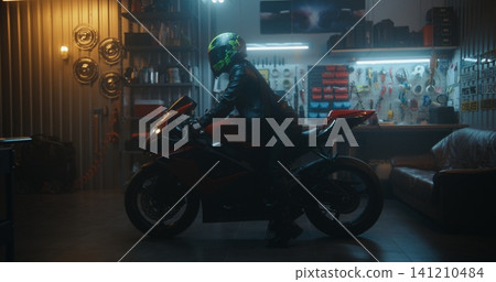Female Biker Putting on Helmet and Starting Motorcycle Engine in Garage Female Biker Putting on Helmet and Starting Motorcycle Engine in Garage 141210484