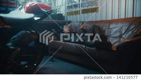 Female Motorcyclist Reclines on Brown Sofa, Using Mobile Device After Ride Female Motorcyclist Reclines on Brown Sofa, Using Mobile Device After Ride 141210577