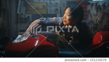 Smiling Female Mechanic Detailing Motorbike With Spray and Cloth on Sunny Day Smiling Female Mechanic Detailing Motorbike With Spray and Cloth on Sunny Day 141210579
