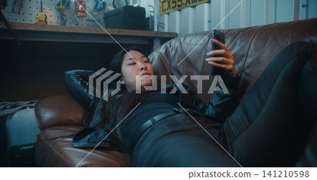 Female Motorcyclist Reclines on Brown Sofa, Using Mobile Device After Ride 141210598